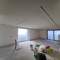 BB-PROBUD - Wnętrze w trakcie remontu: gładkie, białe ściany i sufit z wpuszczonymi liniami oświetleniowymi, okna zabezpieczone folią, widoczne narzędzia i materiały budowlane, przewody elektryczne wystające...