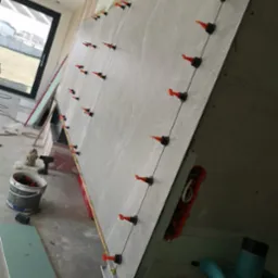 Montaż dużych, szarych płytek na ścianie z użyciem systemu poziomowania z pomarańczowymi klinami, widoczne narzędzia i materiały budowlane, w tle okno i surowy stan wykończenia.