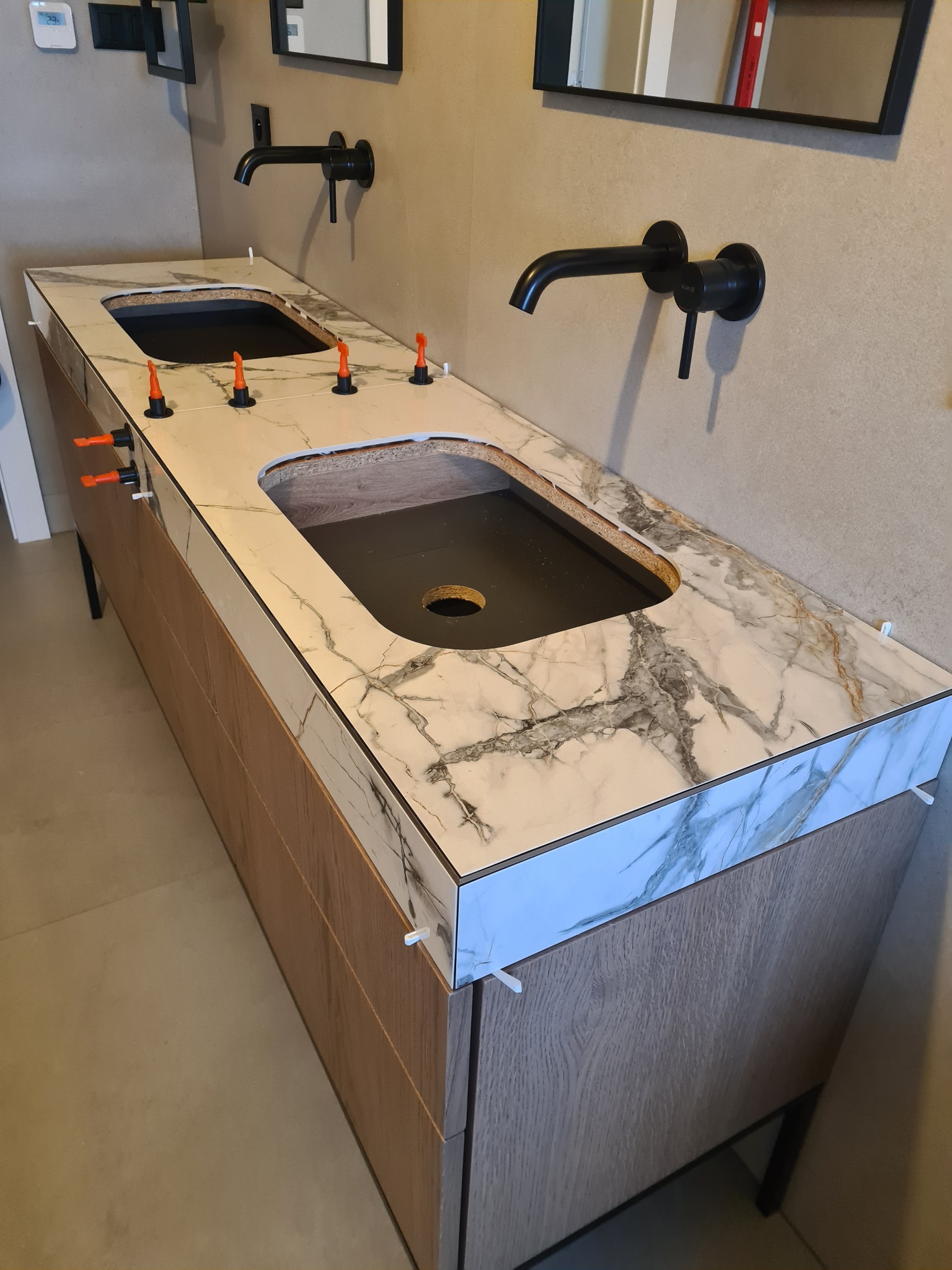 Montaż blatu z płytek imitujących marmur na szafce pod umywalki, widoczne czarne baterie ścienne i system poziomowania płytek.