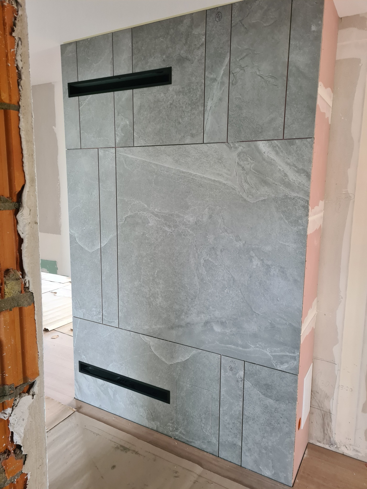 Ściana wykończona płytkami imitującymi kamień w odcieniach szarości z czarnymi wnękami wentylacyjnymi, widoczne surowe wykończenie ścian bocznych z cegły i płyty gipsowej.