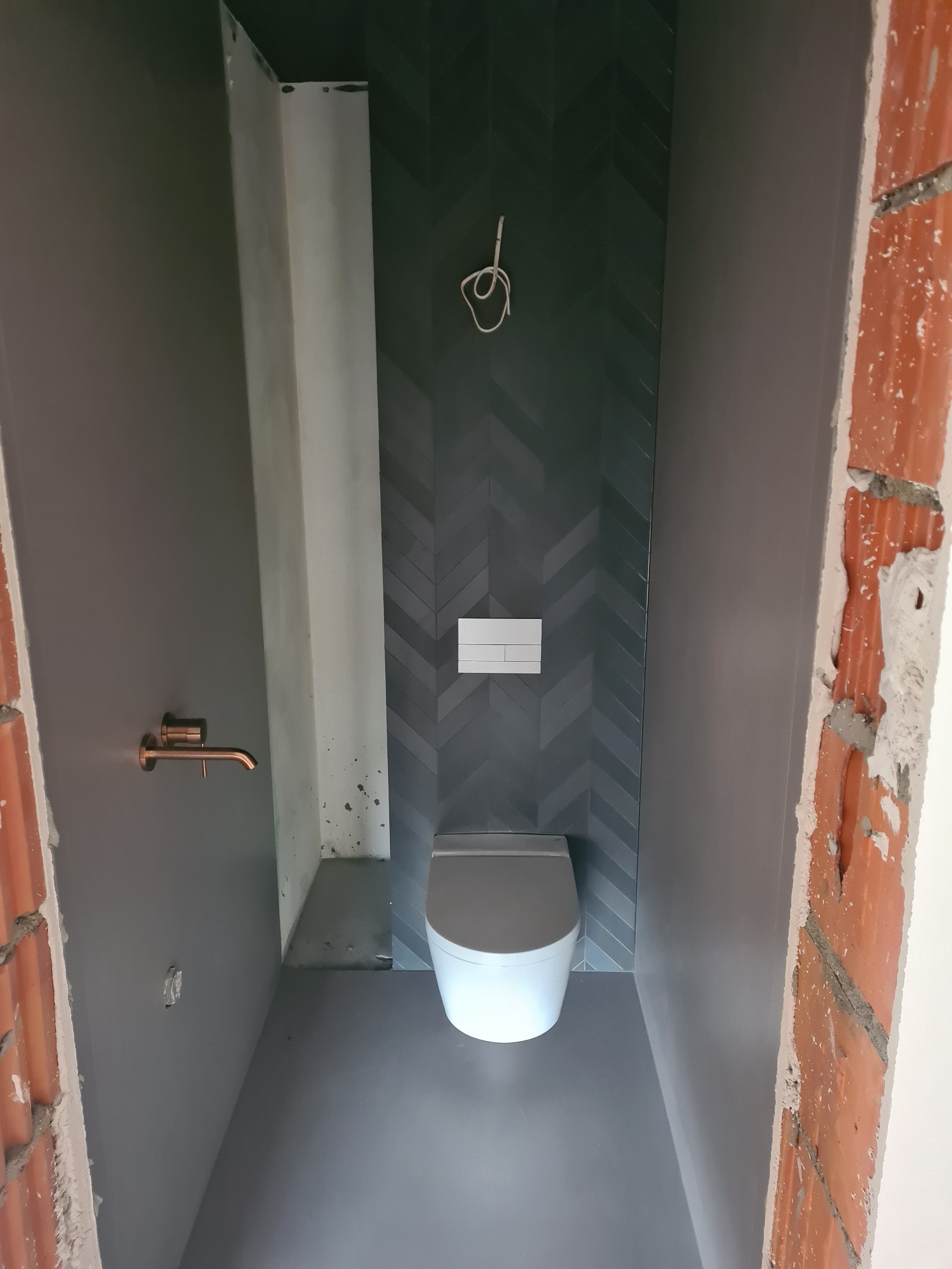 Wąska łazienka w trakcie wykończenia: ciemnoszare ściany, płytki ułożone w jodełkę, biała toaleta, miedziana bateria umywalkowa, widoczne niedokończone obramowania drzwi z cegły.