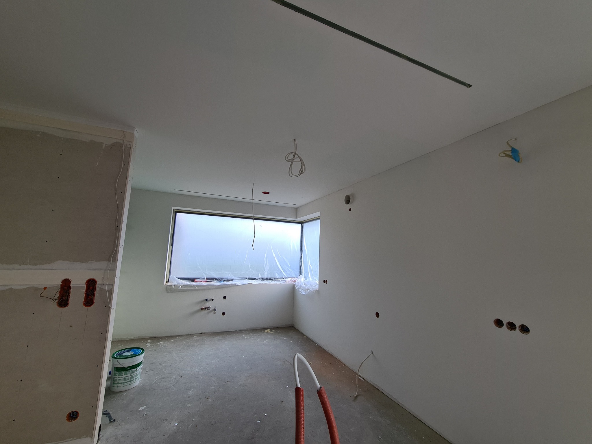 Surowe wnętrze w trakcie prac wykończeniowych: ściany częściowo pokryte płytami gipsowo-kartonowymi, widoczne puszki elektryczne, okno zabezpieczone folią, na podłodze wiadro z farbą i rury...