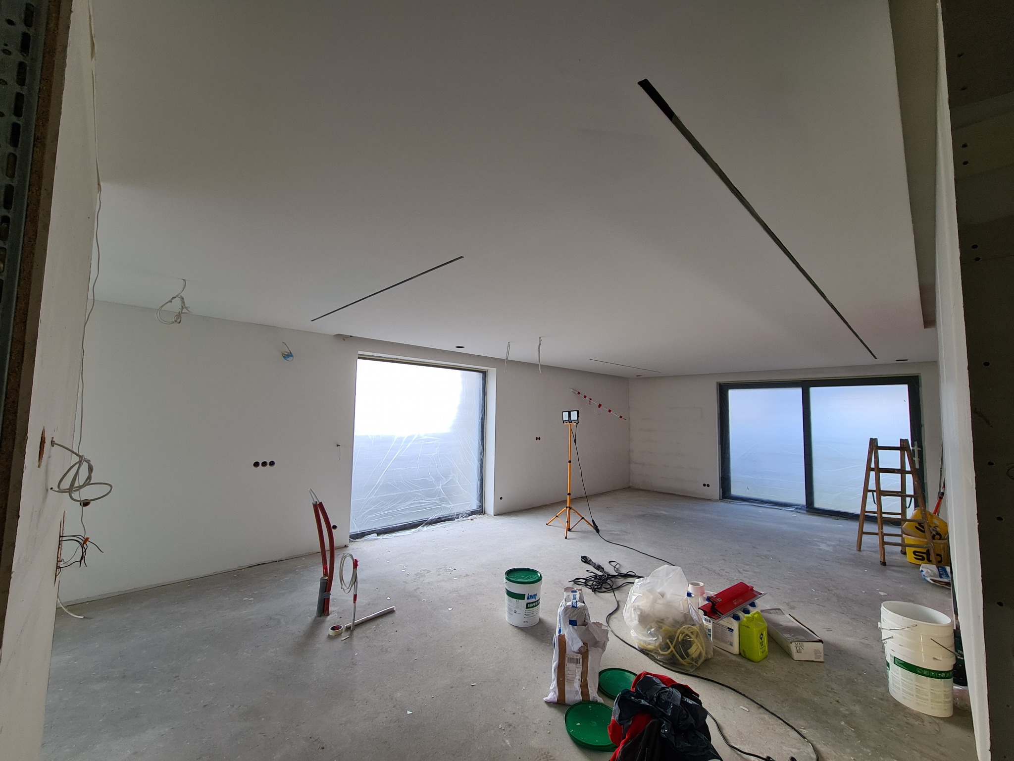 Wnętrze w trakcie remontu: gładkie, białe ściany i sufit z wpuszczonymi liniami oświetleniowymi, okna zabezpieczone folią, widoczne narzędzia i materiały budowlane, przewody elektryczne wystające...