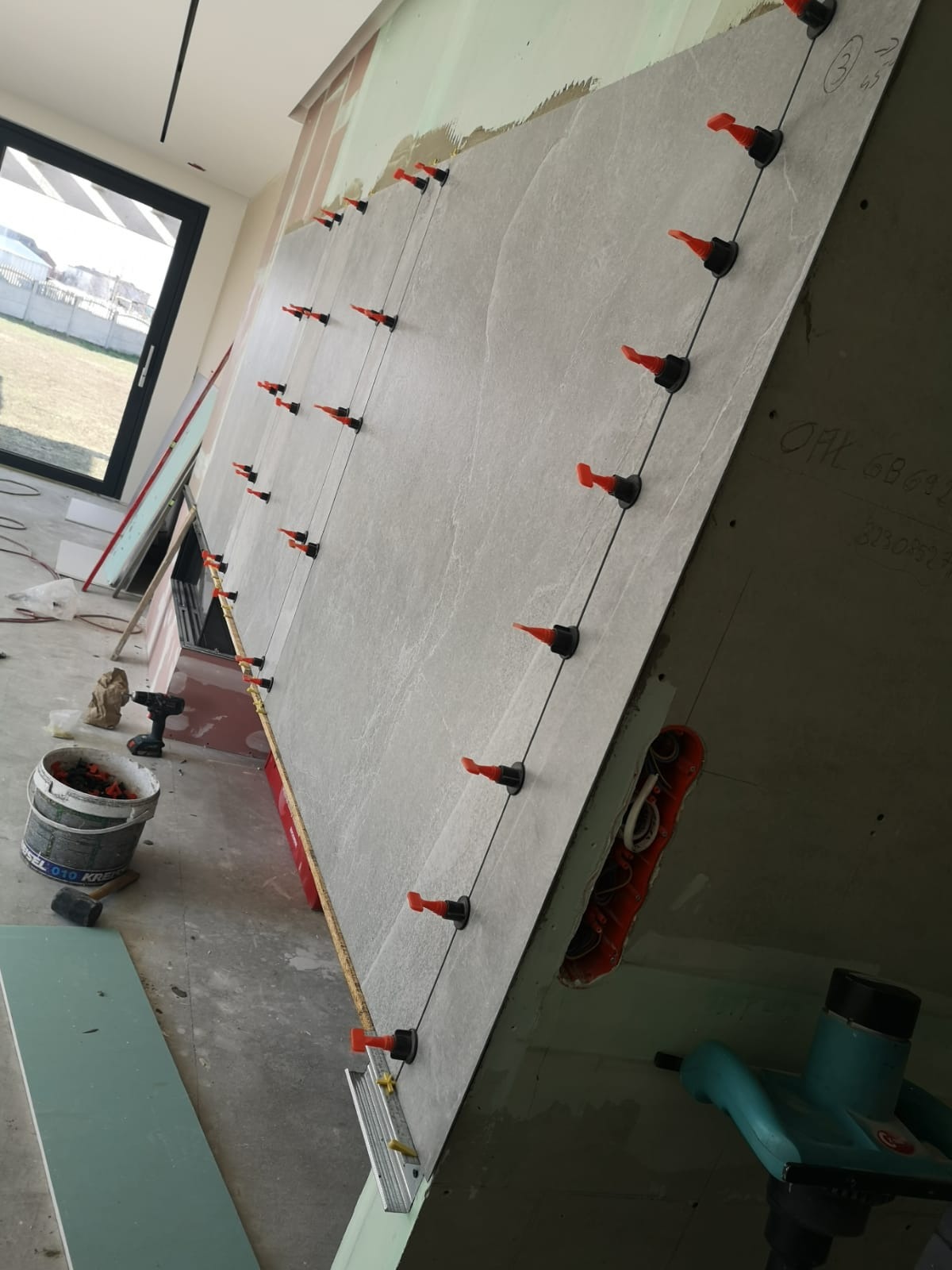 Montaż dużych, szarych płytek na ścianie z użyciem systemu poziomowania z pomarańczowymi klinami, widoczne narzędzia i materiały budowlane, w tle okno i surowy stan wykończenia.