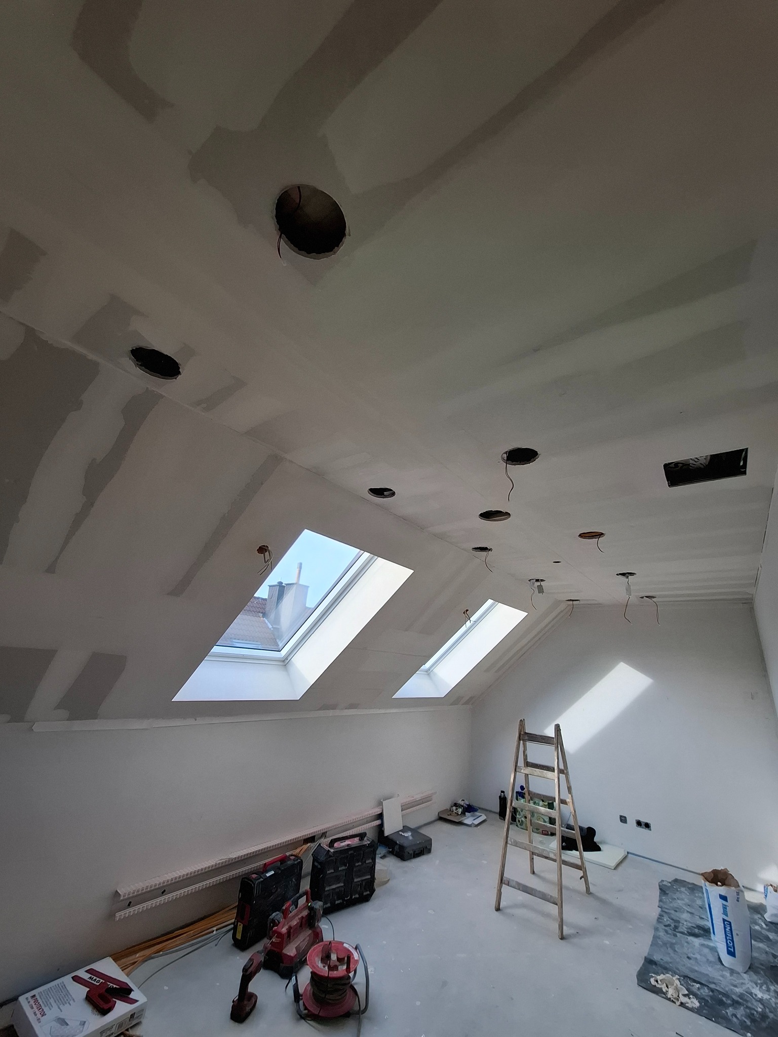 Poddasze w trakcie remontu: widoczne płyty karton-gips na skosach sufitu, otwory na oświetlenie, dwa okna dachowe wpuszczające naturalne światło, drabina i narzędzia na podłodze.