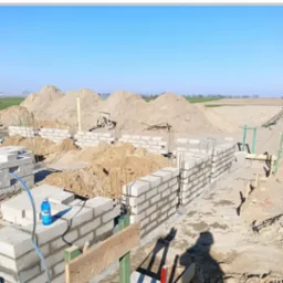 Wznoszenie ścian fundamentowych z bloczków betonowych na placu budowy, widoczne zbrojenia i sterta piasku w tle.