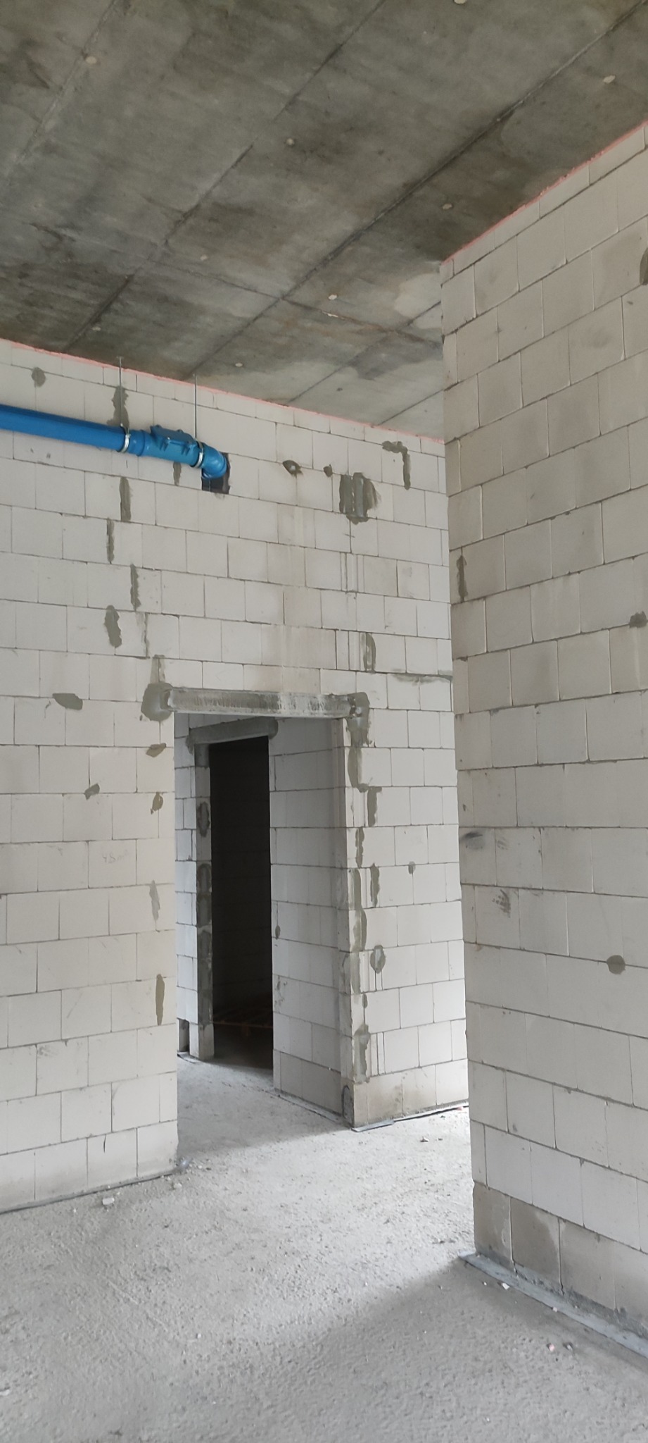 Wnętrze w stanie surowym: ściany z białych bloczków, widoczne niebieskie rury pod sufitem, surowy betonowy strop, otwory drzwiowe bez ościeżnic.