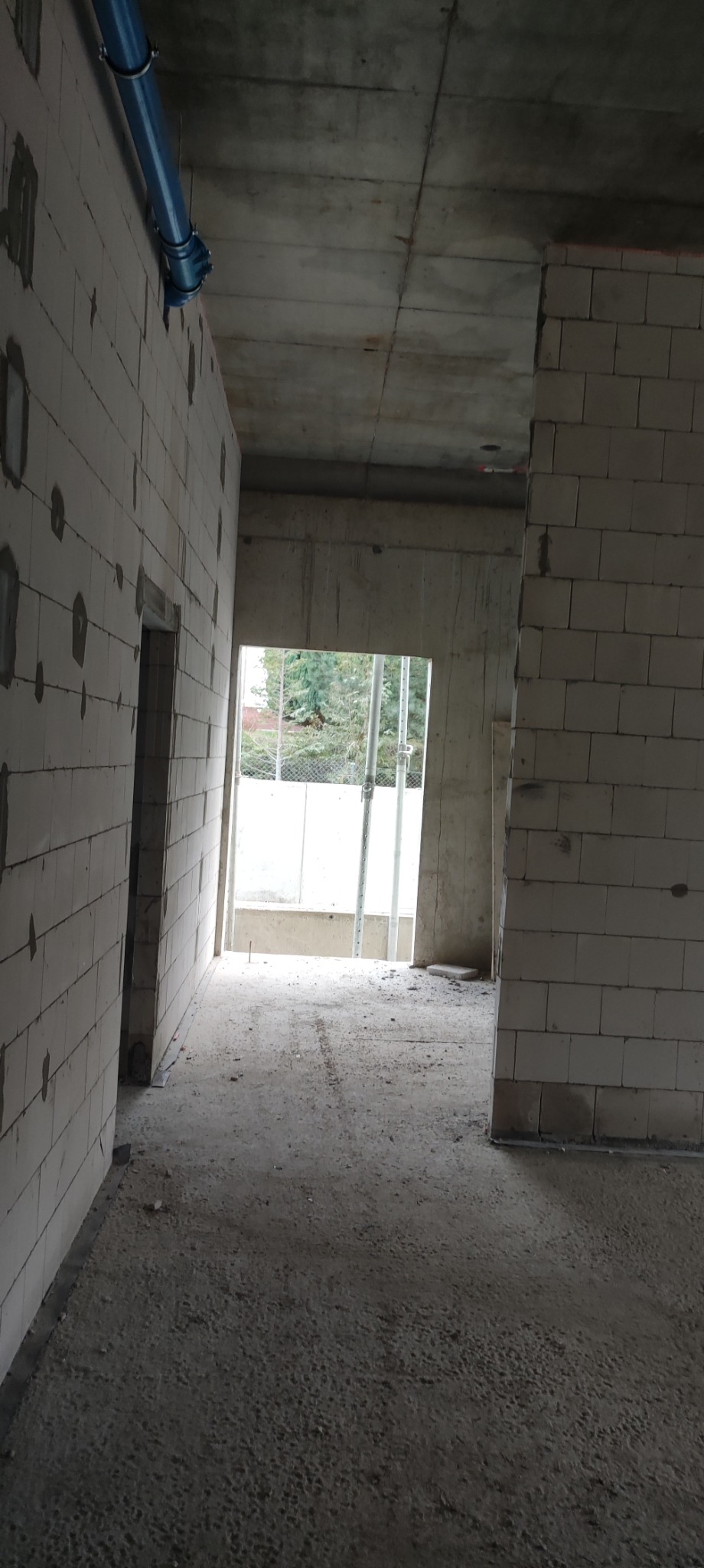 Wnętrze surowego domu w trakcie budowy, widoczne ściany z bloczków, betonowy strop i niebieska rura instalacyjna pod sufitem.