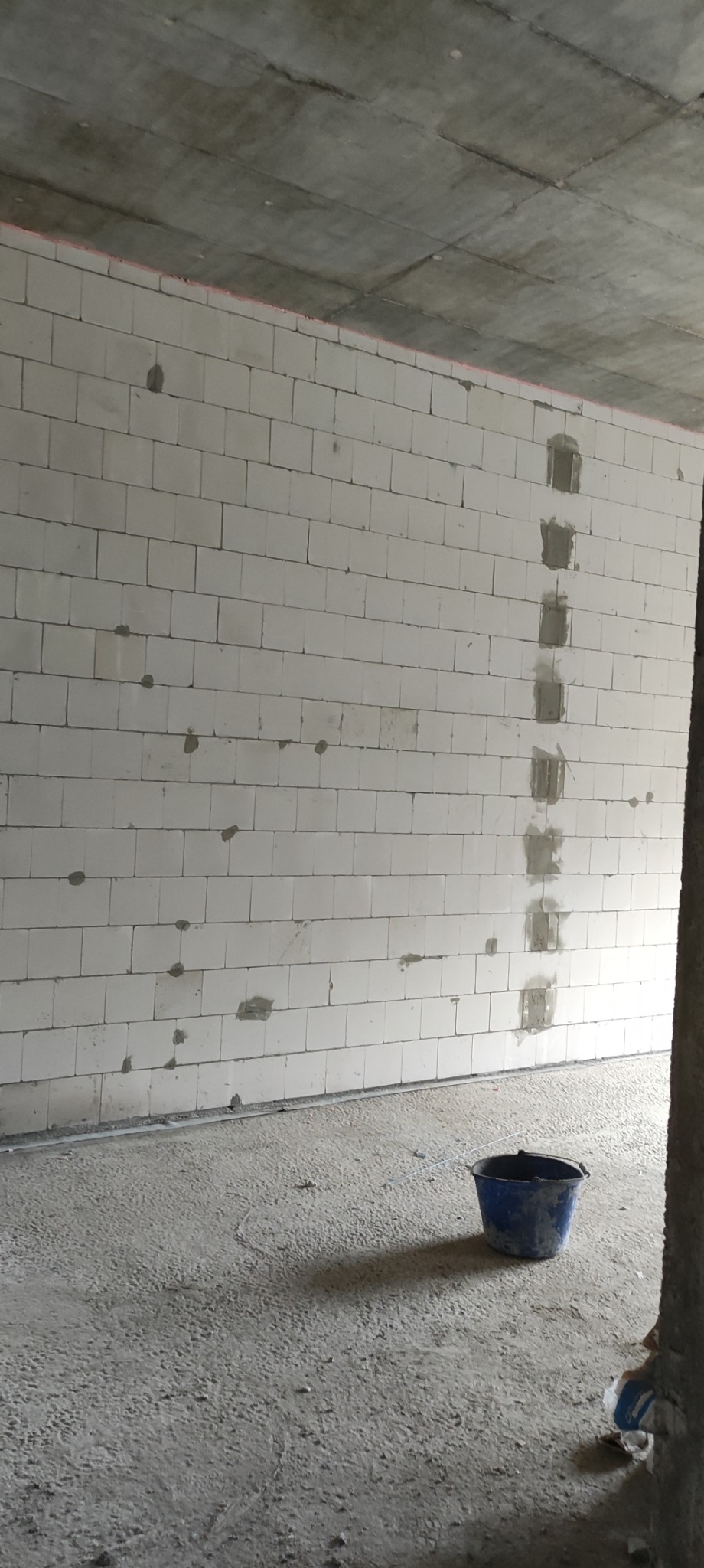 Wnętrze w trakcie budowy: ściana z białych bloczków z wyciętymi otworami na instalacje, surowy betonowy strop, niebieskie wiadro na podłodze.