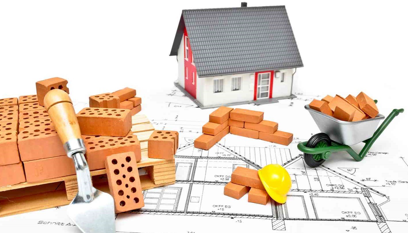 Makieta domu z czerwonymi detalami, cegły, taczka z cegłami, kielnia murarska i żółty kask na planach architektonicznych.