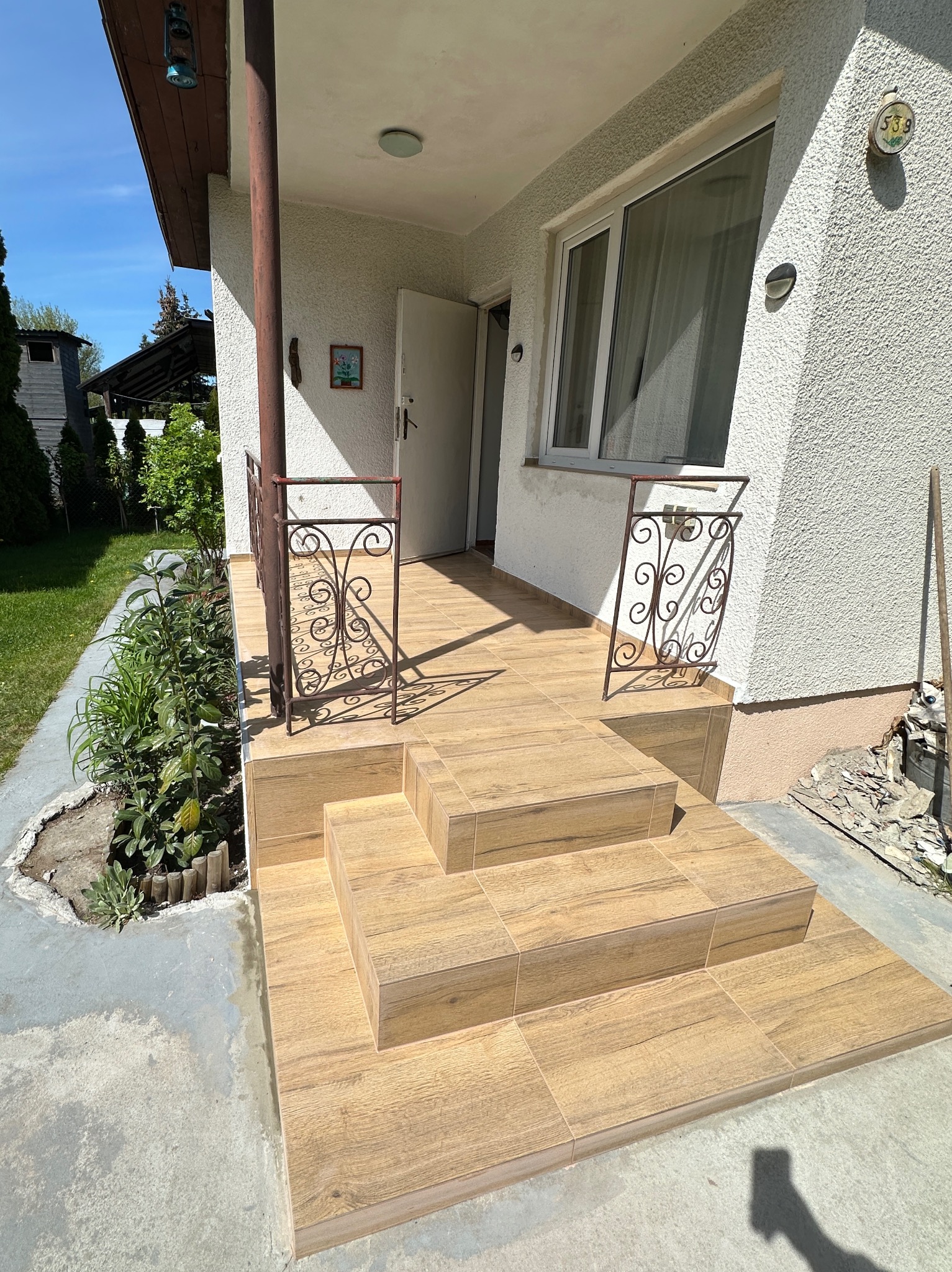 Wykończone płytkami schody wejściowe do domu z ozdobnymi, kutymi barierkami. Płytki imitują drewno. Widoczne świeże nasadzenia roślin obok schodów.