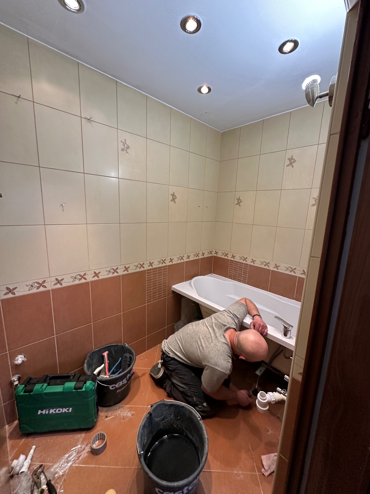 Instalator w trakcie montażu syfonu wannie w łazience z beżowo-brązowymi płytkami. Widoczne narzędzia i akcesoria instalacyjne.