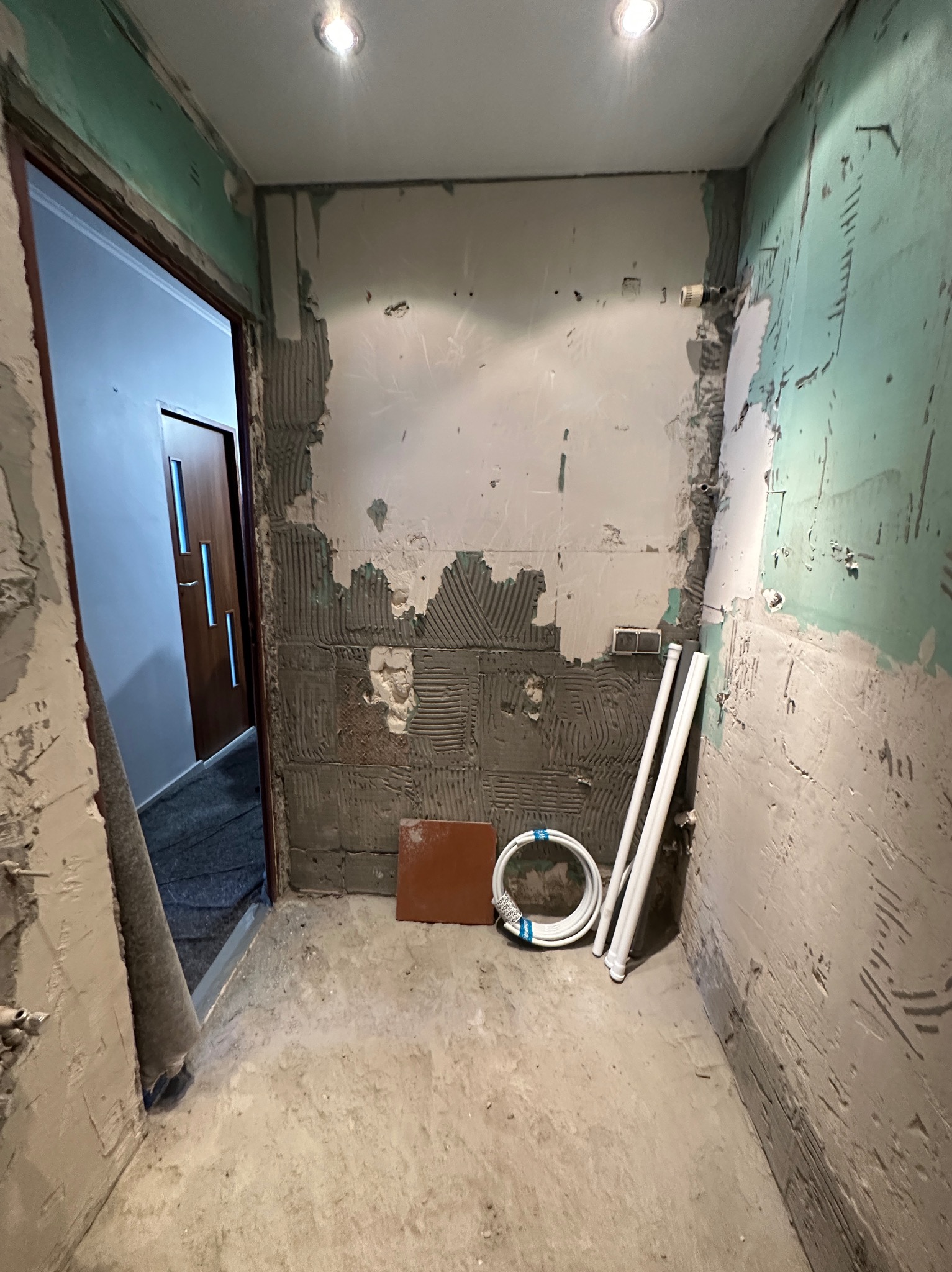 Remont łazienki w Brzozie: ściany przygotowane pod płytki, widoczne rury instalacyjne i pojedyncza płytka. W tle otwarte drzwi do innego pomieszczenia.