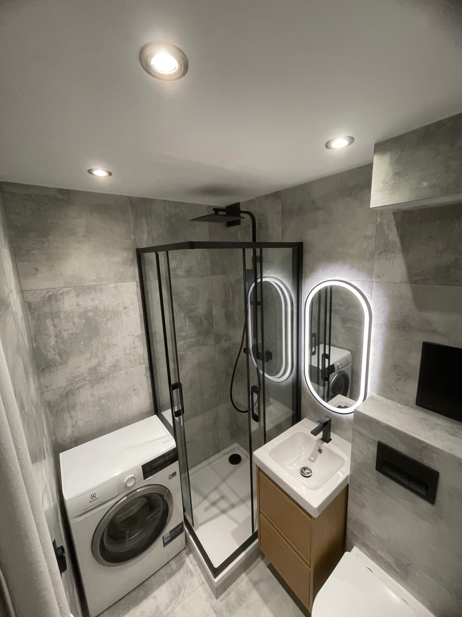 Nowoczesna łazienka w szarościach z kabiną prysznicową typu walk-in, pralką i podświetlanym lustrem. Minimalistyczny design, armatura w czarnym macie.