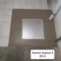 Metalowa klapa włazu wylana betonem na podłodze z płytek, widoczne rury przy ścianie.