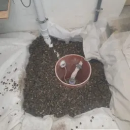 Szczegółowy widok na drenaż wokół rury odpływowej, otoczonej geowłókniną i zasypanej żwirem, z widocznym fragmentem instalacji kanalizacyjnej w tle.