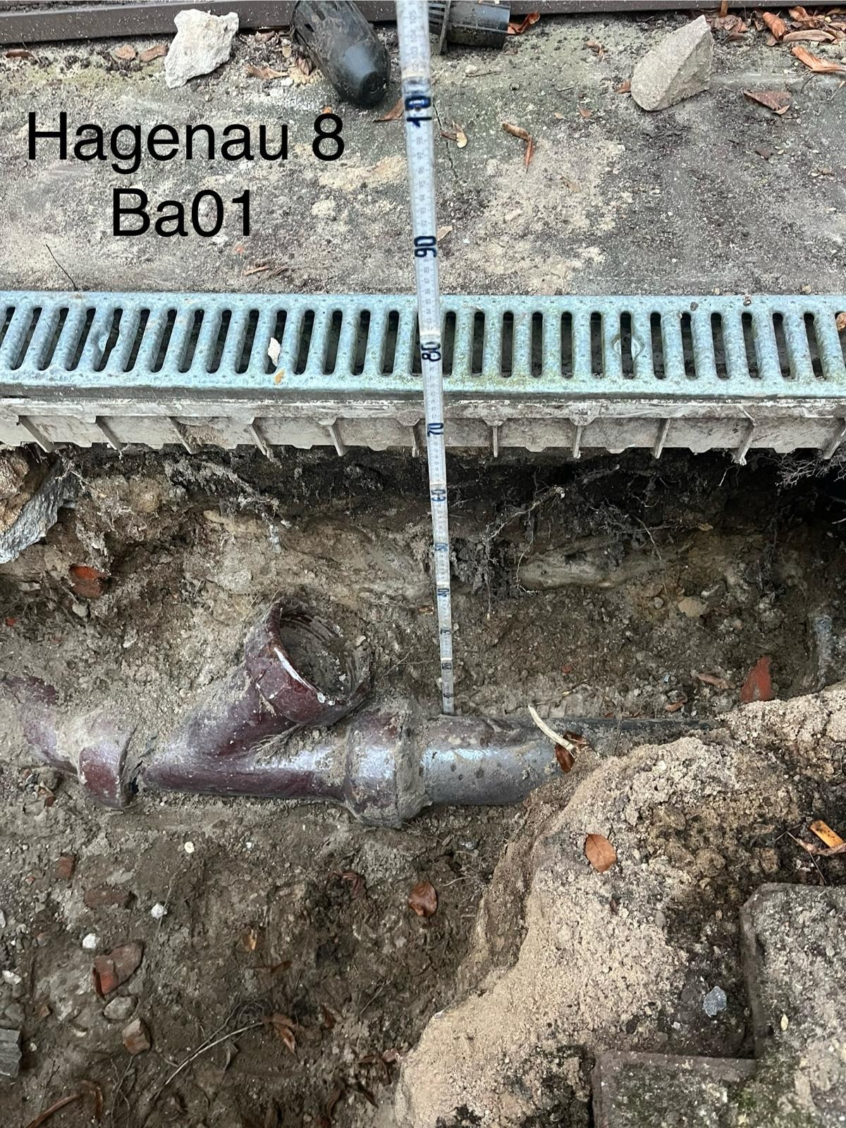 Odkryta instalacja kanalizacyjna w ziemi, widoczne rury łączące się w kształt litery Y, pomiar głębokości wykopu za pomocą miarki, metalowa kratka ściekowa w tle, ziemia z gruzem.