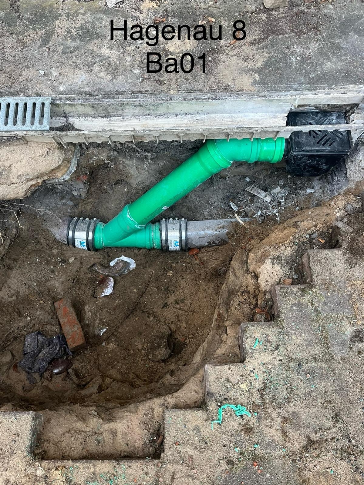 Połączenie rur kanalizacyjnych: zielona rura z trójnikiem i złączkami w wykopie ziemnym obok betonowych schodów i kratki ściekowej.