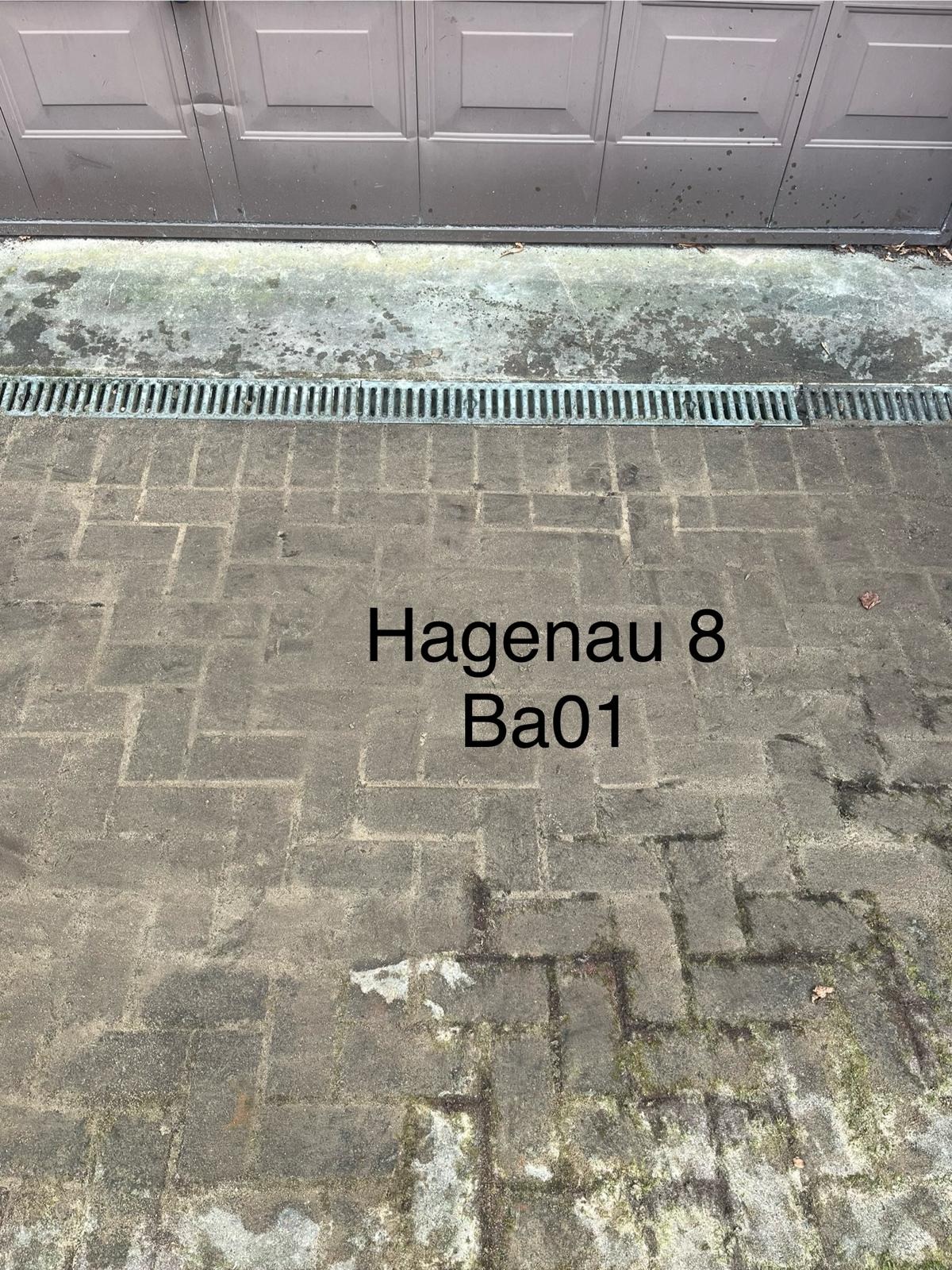 Brudna kostka brukowa przed garażem z widoczną kratką odpływową i napisem 'Hagenau 8 Ba01'.