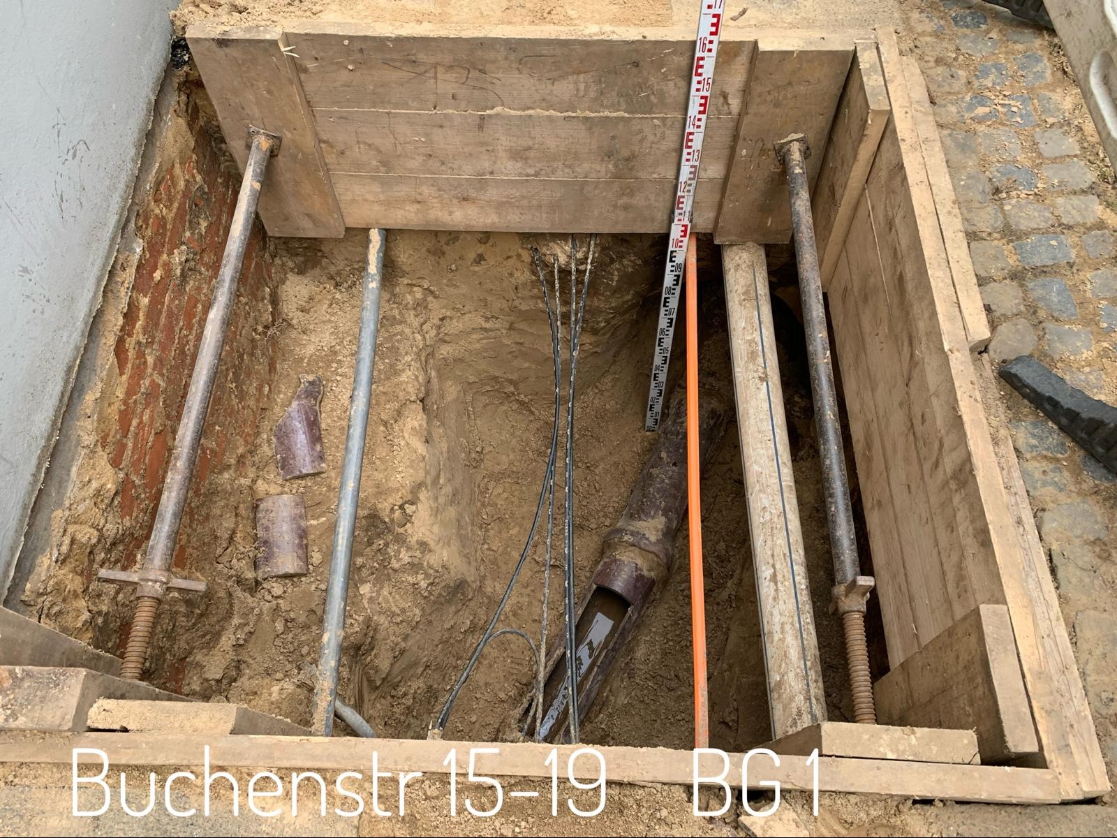 Wykop zabezpieczony drewnianą szalunką i stalowymi rozpórkami, widoczna rura kanalizacyjna w ziemi, przymiar geodezyjny oparty o ściankę wykopu.