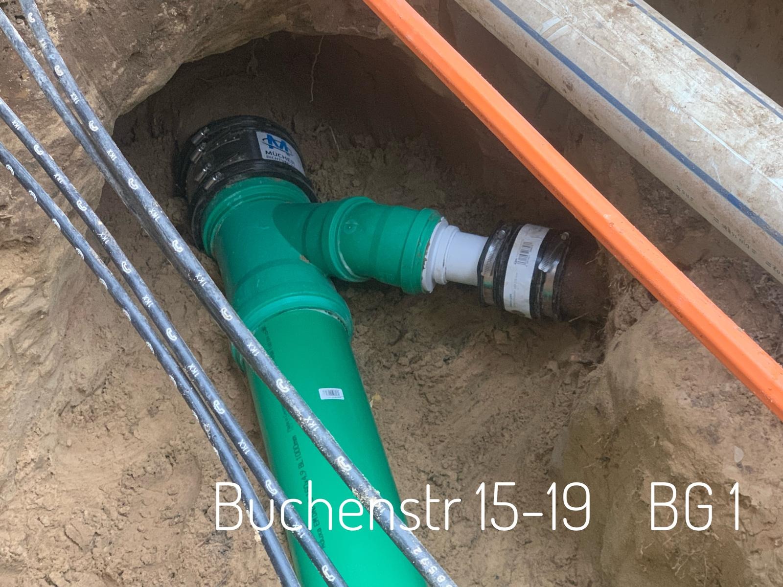 Zielona rura kanalizacyjna z łącznikiem w wykopie, widoczne kable i inne rury, ziemia wokół.