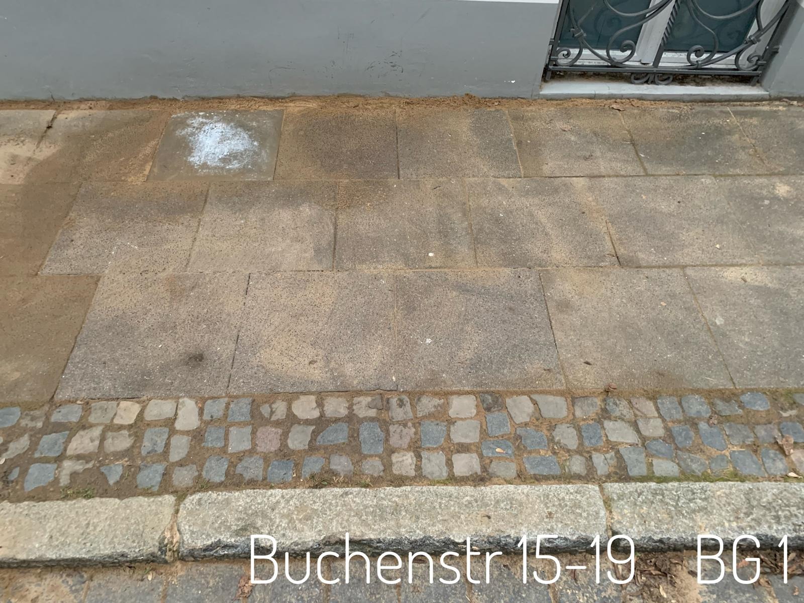 Ułożona kostka brukowa z piaskiem na powierzchni, fragment chodnika przy budynku z widocznym adresem Buchenstr 15-19 BG1