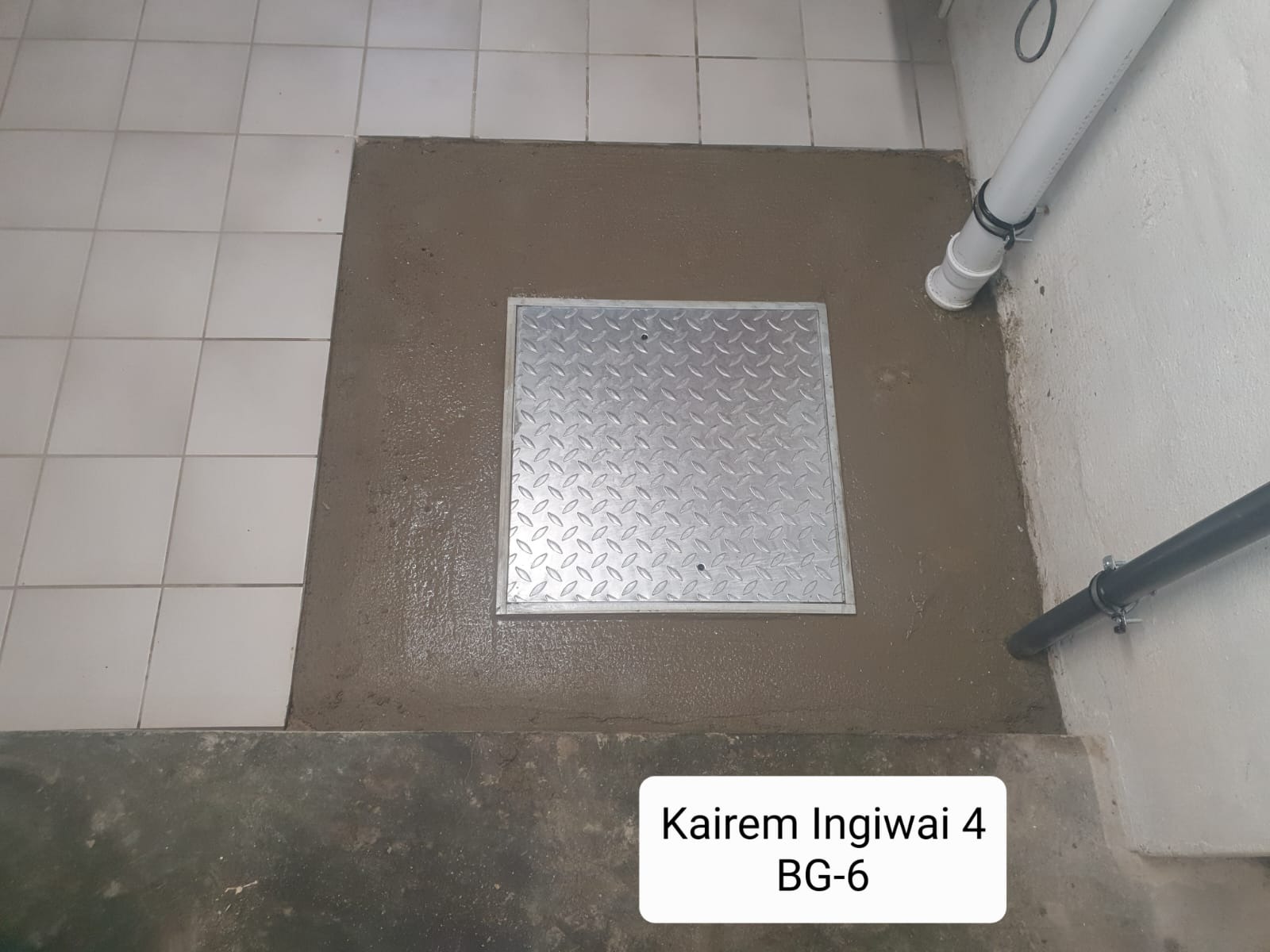 Metalowa klapa włazu wylana betonem na podłodze z płytek, widoczne rury przy ścianie.