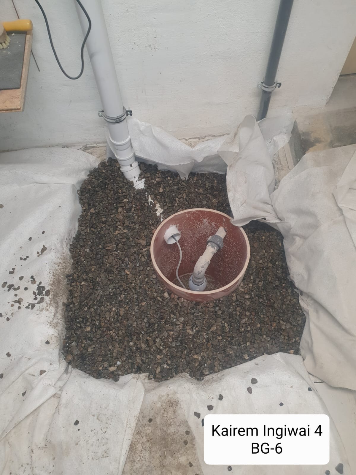 Szczegółowy widok na drenaż wokół rury odpływowej, otoczonej geowłókniną i zasypanej żwirem, z widocznym fragmentem instalacji kanalizacyjnej w tle.