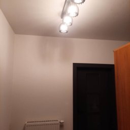 Paweł Walczak - Wnętrze pomalowanego na jasny odcień przedpokoju z grzejnikiem, czarnymi drzwiami i lampą sufitową z czterema kloszami