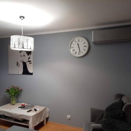 Paweł Walczak - Szara ściana w salonie po malowaniu, z wiszącym zegarem, klimatyzatorem, obrazem Audrey Hepburn i lampą z motywem drzew.