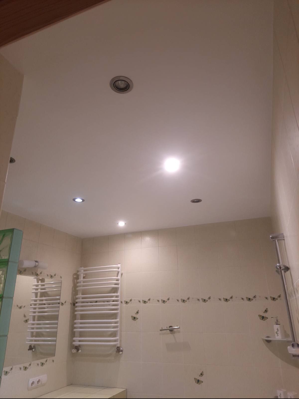 Wykończona łazienka z kremowymi płytkami zdobionymi motylami, grzejnikiem drabinkowym i równo pomalowanym sufitem z czterema punktowymi lampami.