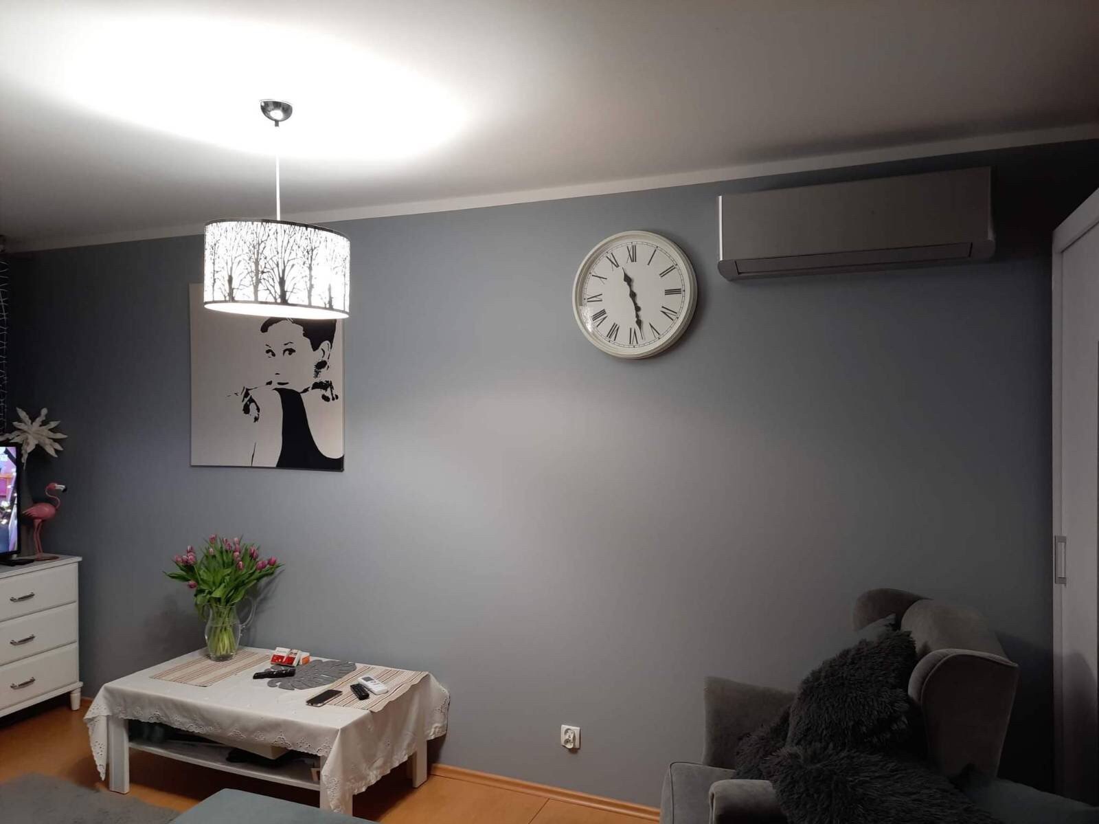 Szara ściana w salonie po malowaniu, z wiszącym zegarem, klimatyzatorem, obrazem Audrey Hepburn i lampą z motywem drzew.
