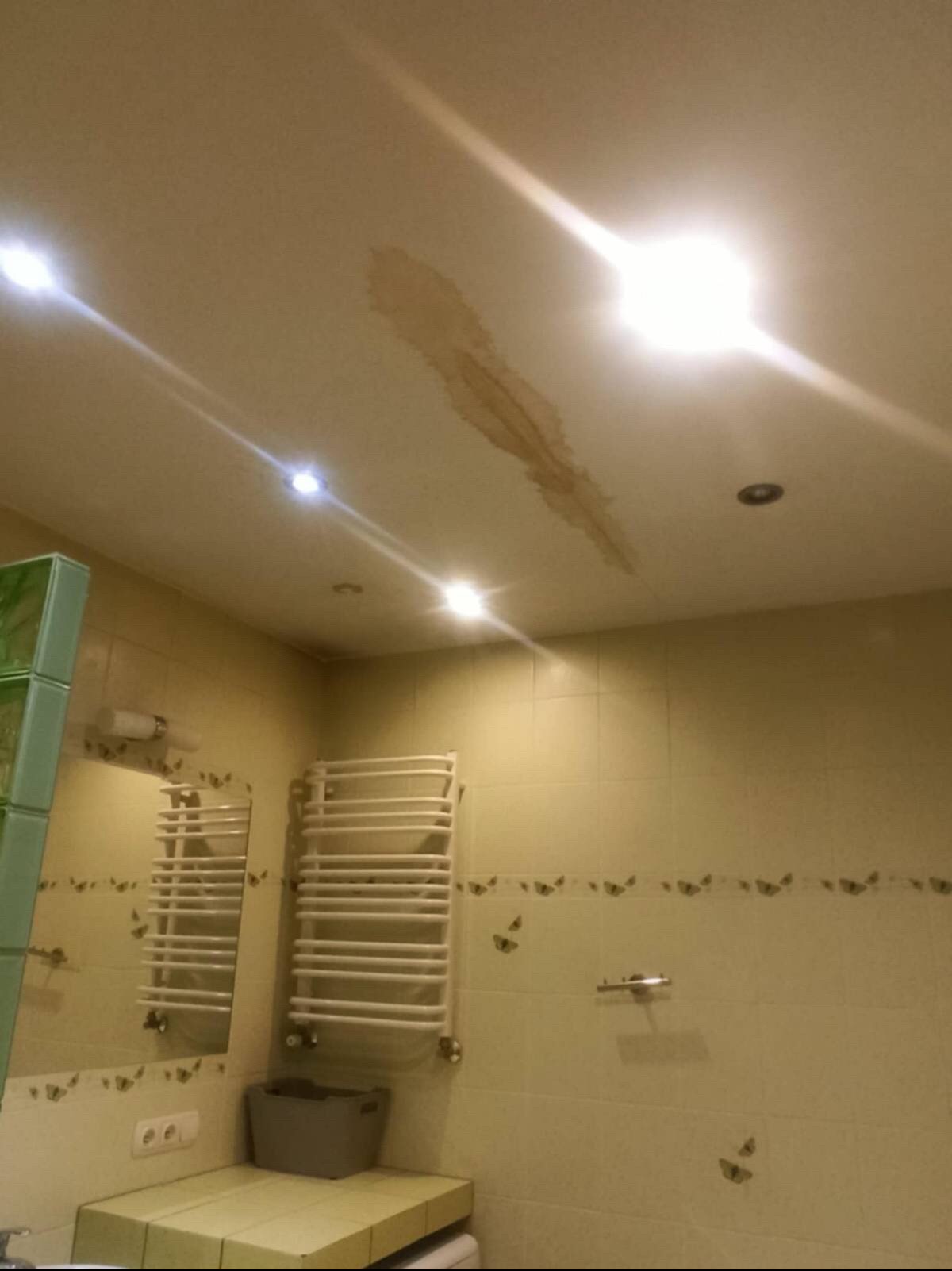 Brązowy zaciek na suficie w łazience z widocznymi punktowymi lampami i fragmentem grzejnika drabinkowego na ścianie w tle.