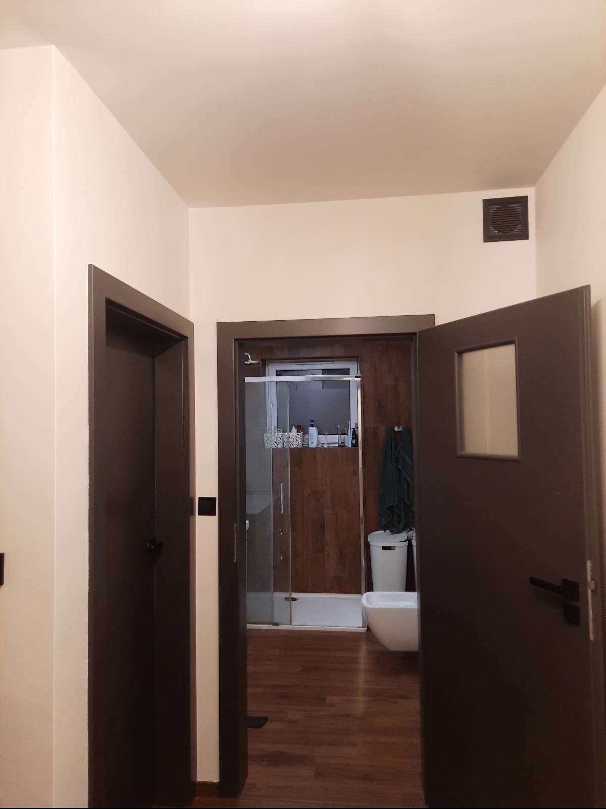 Widok na łazienkę z kabiną prysznicową i toaletą przez otwarte, ciemne drzwi z kwadratowym okienkiem, ściany pomalowane na jasny beż.