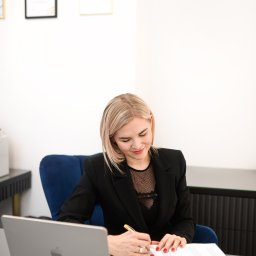 KANCELARIA BANKOWA KINGA BOYKE - Blondynka w czarnej marynarce pisze w notatniku przy biurku z laptopem. Na ścianie wiszą dyplomy.