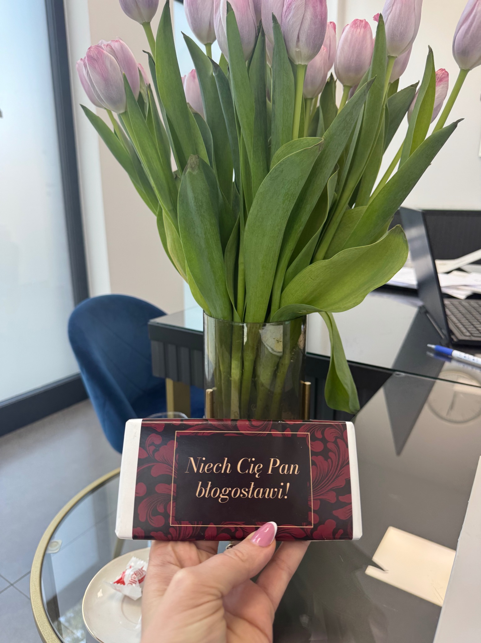 Biuro doradcy: bukiet tulipanów w wazonie, czekolada z napisem 'Niech Cię Pan błogosławi!', filiżanka i nowoczesne biurko z laptopem w tle.