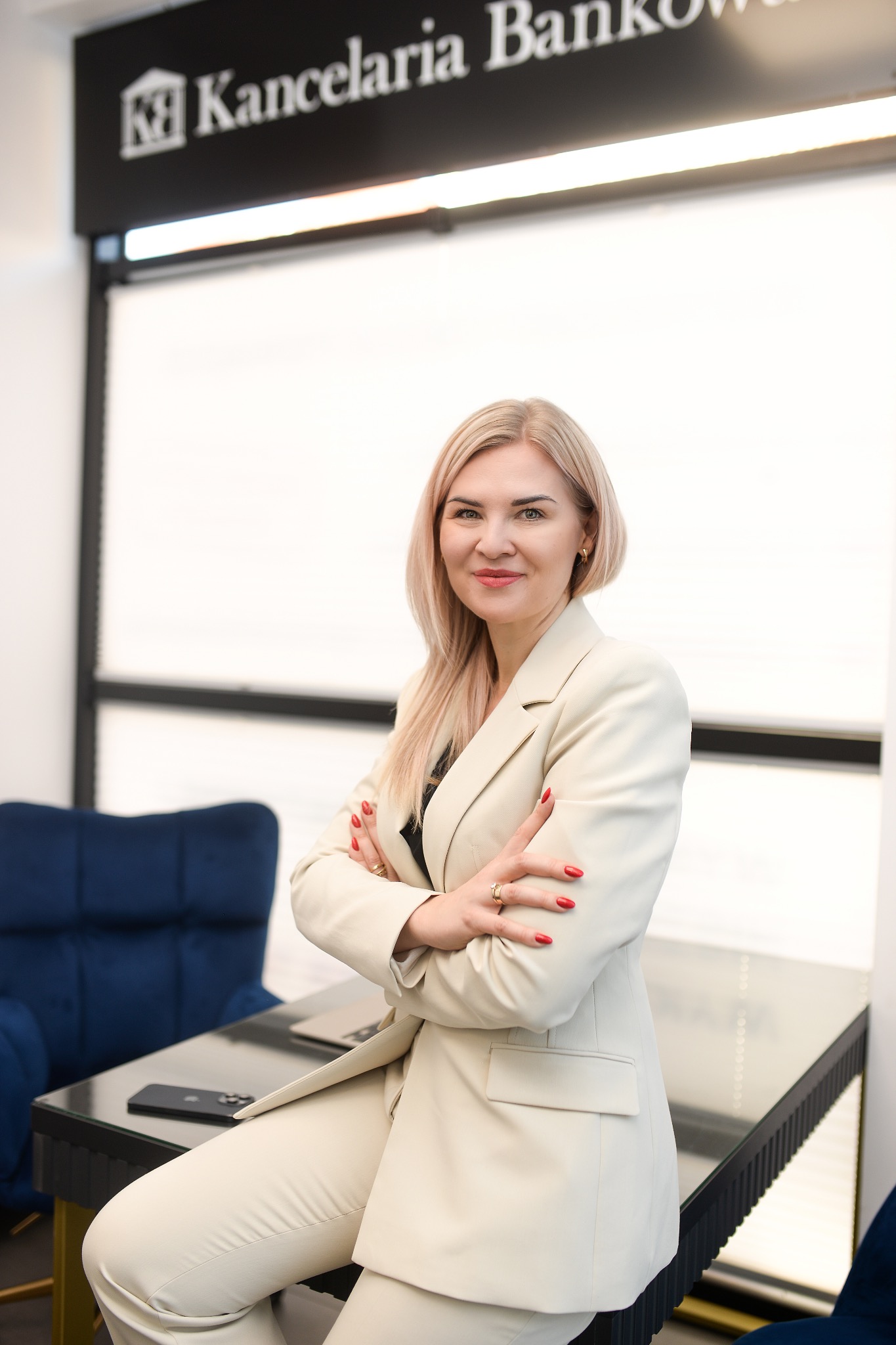 Portret kobiety w jasnym garniturze, z założonymi rękami, siedzącej na biurku w biurze. W tle logo 'Kancelaria Bankowa' i niebieski fotel.