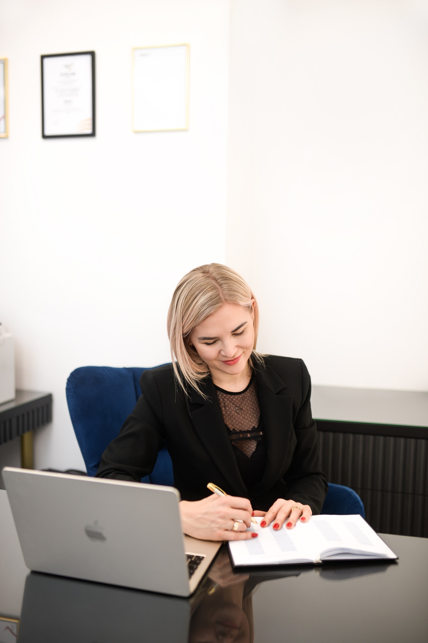 Blondynka w czarnej marynarce pisze w notatniku przy biurku z laptopem. Na ścianie wiszą dyplomy.