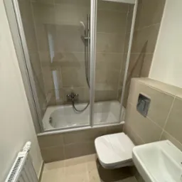 Beżowa łazienka z kabiną prysznicową nad wanną, wiszącą toaletą, umywalką i grzejnikiem żeberkowym; widoczna armatura i płytki w odcieniach beżu.