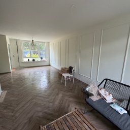 B.K.Montage - Pokój po remoncie z parkietem ułożonym w jodełkę, białe ściany z dekoracyjnymi listwami, meble tymczasowo ustawione w pomieszczeniu, widok na okno z zielenią za nim.