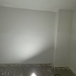 B.K.Montage - Świeżo pomalowane na biało ściany w pokoju, zabezpieczona taśmą malarską podłoga, lampa robocza na podłodze oświetla pomieszczenie
