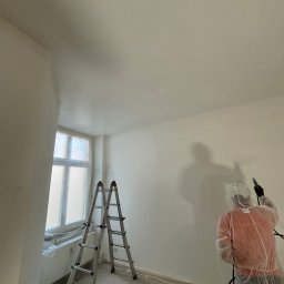 B.K.Montage - Pracownik w kombinezonie ochronnym maluje ścianę natryskowo w pomieszczeniu z zabezpieczonym oknem i rozstawioną drabiną.