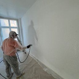 B.K.Montage - Pracownik w kombinezonie ochronnym maluje ścianę natryskowo na biało, widoczna drabina i folia zabezpieczająca podłogę.