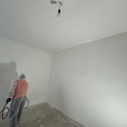 B.K.Montage - Pracownik w kombinezonie malarskim natryskuje białą farbę na ścianę w pokoju z gołą żarówką na suficie, widoczne ślady farby na podłodze zabezpieczonej folią.