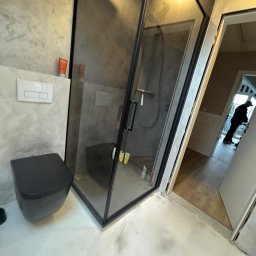 B.K.Montage - Nowoczesna łazienka z czarną armaturą: kabina prysznicowa walk-in z grafitowymi płytkami, wisząca toaleta z ukrytym systemem spłukiwania i biała posadzka z żywicy epoksydowej. W tle widoczna osoba...