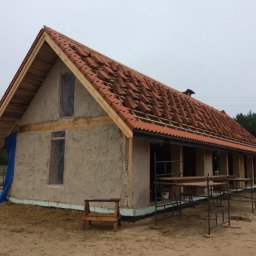 B.K.Montage - Budowa domu z gliny z częściowo ułożoną czerwoną dachówką, okna zabezpieczone folią, widoczne rusztowania i konstrukcja drewniana dachu.