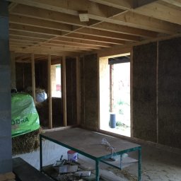 B.K.Montage - Wnętrze domu w trakcie budowy z bali słomy, widoczne ściany z wypełnieniem słomą, drewniana konstrukcja stropu, worki ze słomą przy ścianie, stół roboczy z narzędziami.