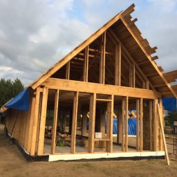 B.K.Montage - Surowa konstrukcja domu z drewna, z widocznymi belami słomy w ścianach, zabezpieczona niebieską plandeką, w tle zieleń drzew i pochmurne niebo.