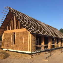 B.K.Montage - Surowy stan domu ze słomy z drewnianą konstrukcją dachu, widoczne rusztowania i bale słomy na ziemi.