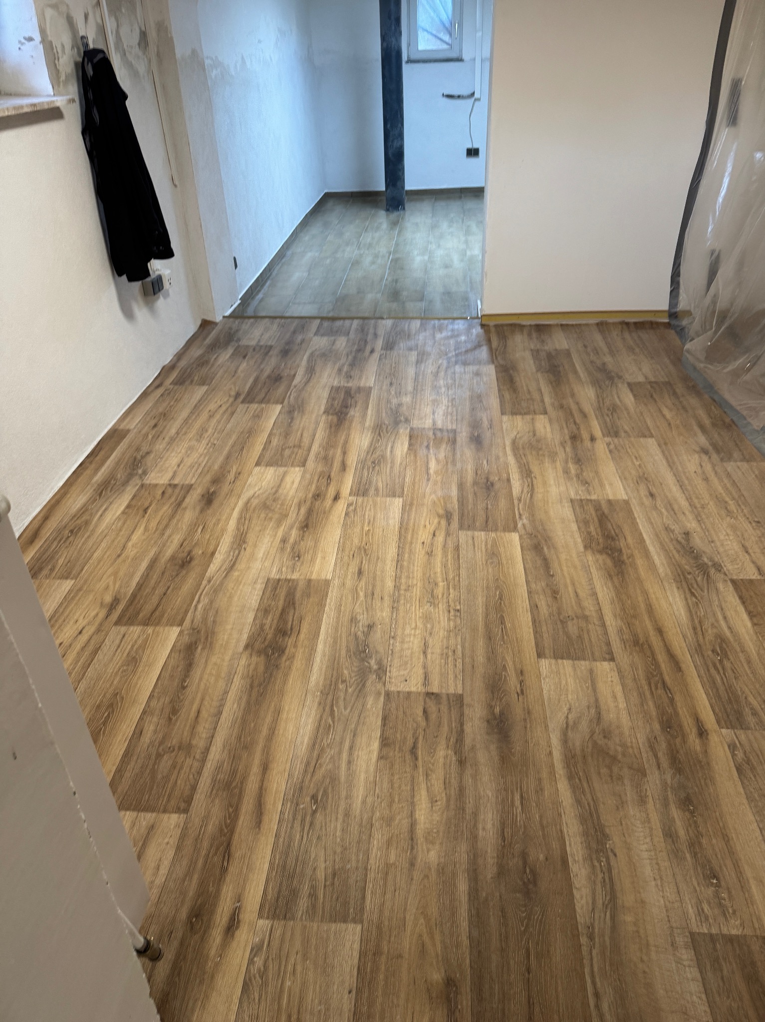 Ułożona podłoga z paneli imitujących drewno w pomieszczeniu w trakcie prac wykończeniowych, widoczne zabezpieczenia folią malarską i wisząca kurtka.